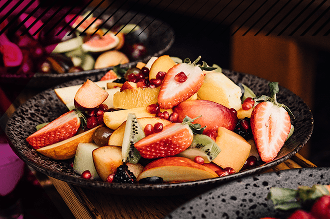 A bowl of fruit prepared with strawberries, kiwi, apple, oranges, peaches, garnished with pomegranate seeds.