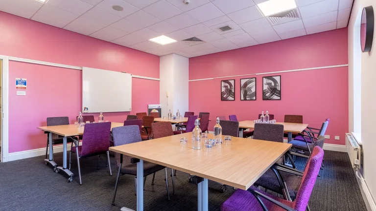 Meeting room with four tables surrounded by chairs with bottles of water and glasses on each table.  Room has pink walls