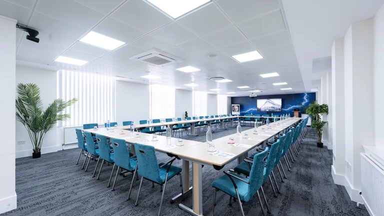 Large meeting room with several blue desk chairs and tables in a square shape with palm trees 