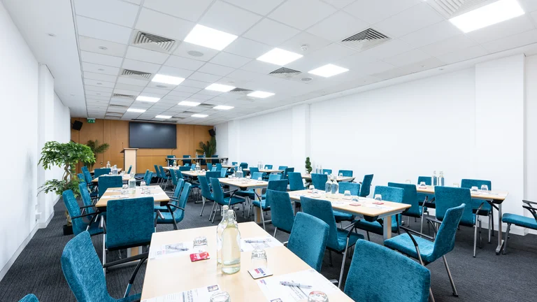Medium Meeting room size with multiple desks surrounded by blue chairs with screen on far wall