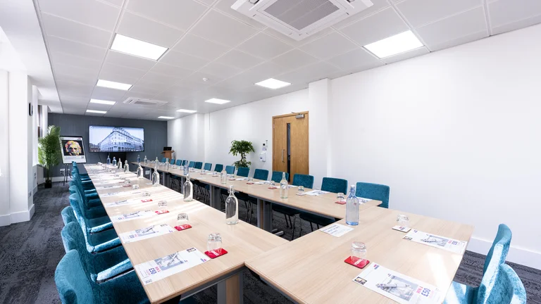 Medium meeting room with blue chairs with lead desk. Screen on wall for presenting