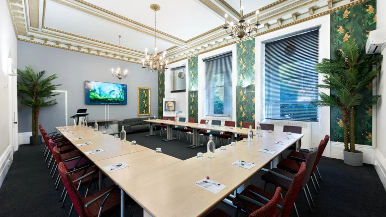 Medium sized meeting room with U shape desk set up surrounded by chairs with screens on the TV and palm trees with tropical design on the walls