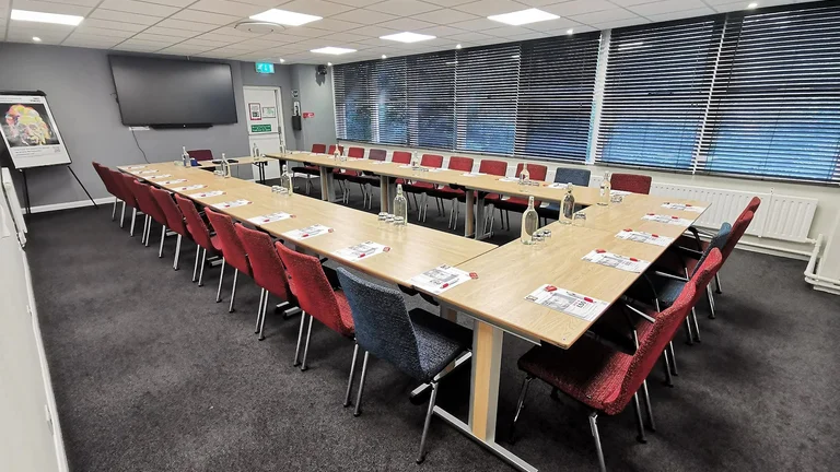 Medium sized meeting room with U shape set up of desks and chairs. Screen on the far wall for presenting.