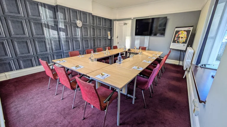 Medium Meeting room with rectangular set up of desks and chairs with large screen for conferencing and a board in the corner for flipcharts. Panel print on the walls.