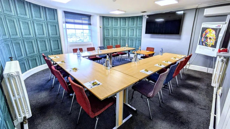 Meeting room with u shaped desks, tv screen and panelling print on the wall