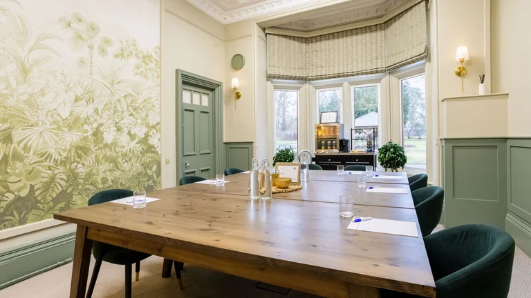 Executive suite at Woodland Grange featuring a central table surrounded by chairs,  with two gass bottles of water placed neatly on the tabletop , fridge, large window and leaf wallpaper design on walls 