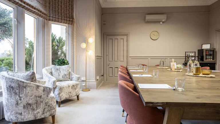 Two armchairs sit by large glass windows, large table in the centre surrounded by chairs with tea station in corner