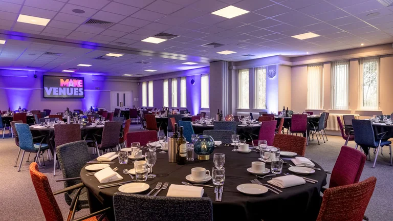 Large conference room with several circular tables with place settings and colourful chairs, with a TV screen at the front of the room