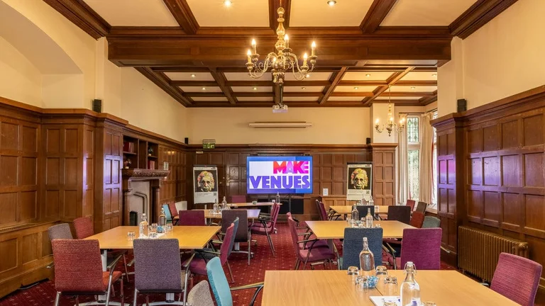 A large meeting room with wood-panelled walls, a coffered ceiling, and chandelier lighting,  featuring multiple rectangular tables with colourful chairs, glass bottles of water,  and a large tv screen displaying the "Make Venues" logo.