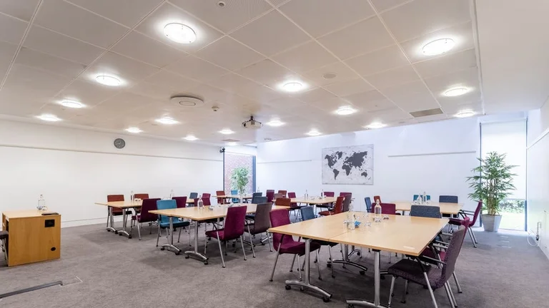 Meeting room with several rectangular tables surrounded by chairs; a single-person desk at the front for presentations, and a world map print on the wall