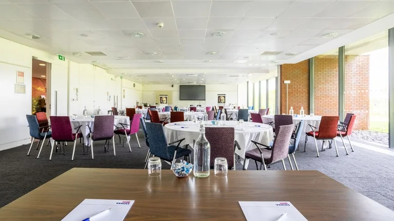 Large meeting room with multiple circular tables surrounded by colourful chairs, with a large TV screen at the front of the room and floor to ceiling windows allowing natural light in, the forefront of the image is a desk with water glasses
